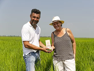 Mann mit Frau in sommerlichr Kleidung auf einem Reisfeld. Beide halten gemeinsam eine Packung Rapunzel Risotto