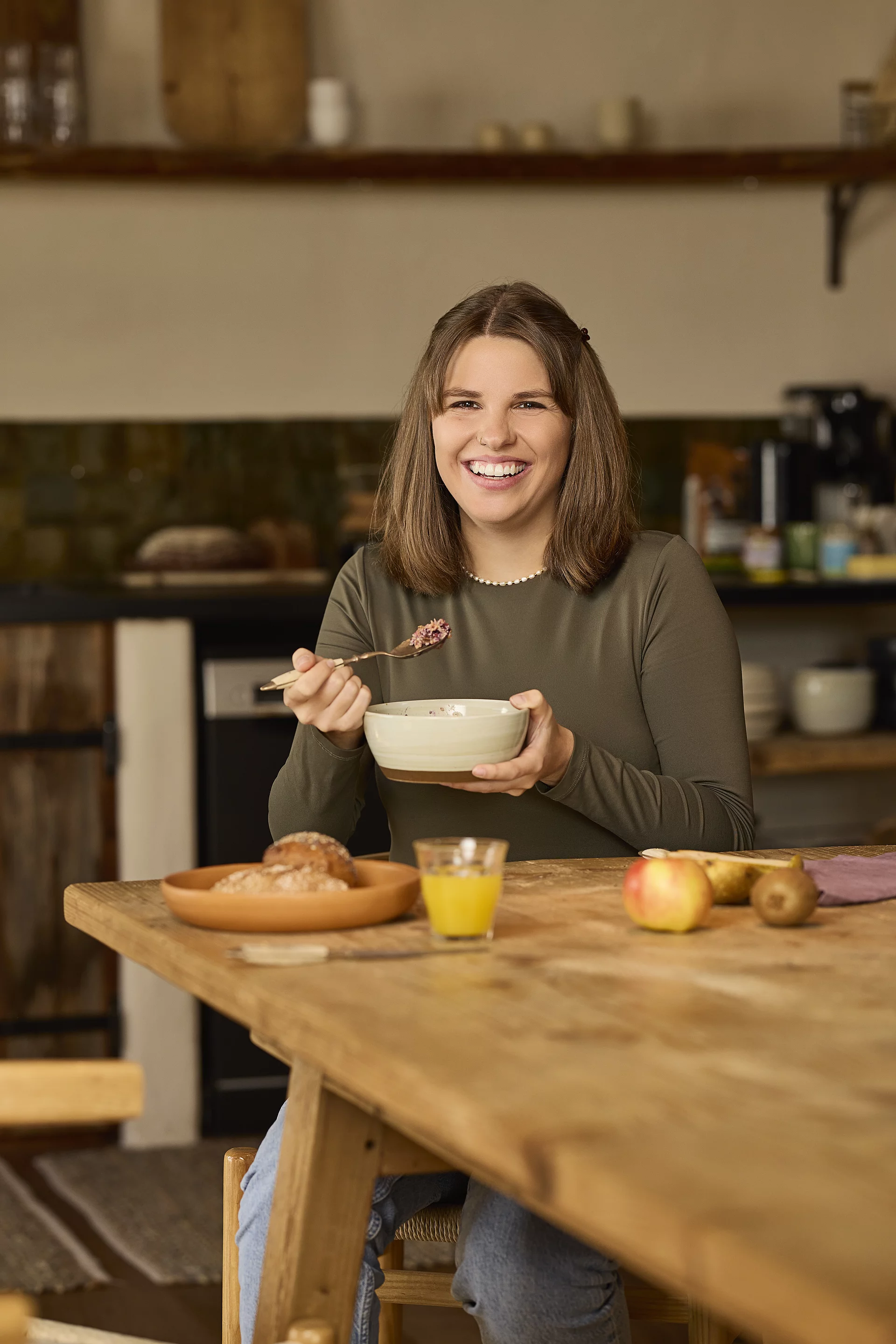 Genussvoll in den Tag mit Beerenmüsli. Eine junge Frau sitzt lächelnd am Frühstückstisch und hält eine Schale Beerenmüsli in der Hand. Auf dem Tisch stehen frische Brötchen und ein Glas frisch gepresster Orangensaft.