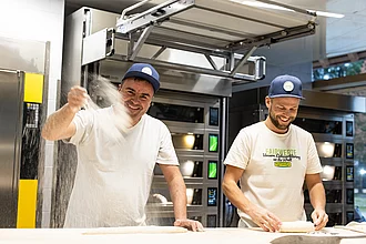 Zwei Bäcker am Backtisch in der Rapunzel Bäckerei bei der Arbeit, einer wirft Mehl in die Luft, beide lachen.