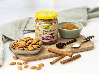 Glas Rapunzel Retromus Gebrannte Mandel auf Küchenoberfläche, daneben die Zutaten wie Mandeln und Rohrzucker – wenige, natürliche Bio-Zutaten für authentischen Genuss