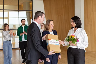 Drei Menschen stehen im Vordergrund. Es wird ein Rapunzel Dankespaket überreicht. Im Hintergrund zwei klatschende Menschen.