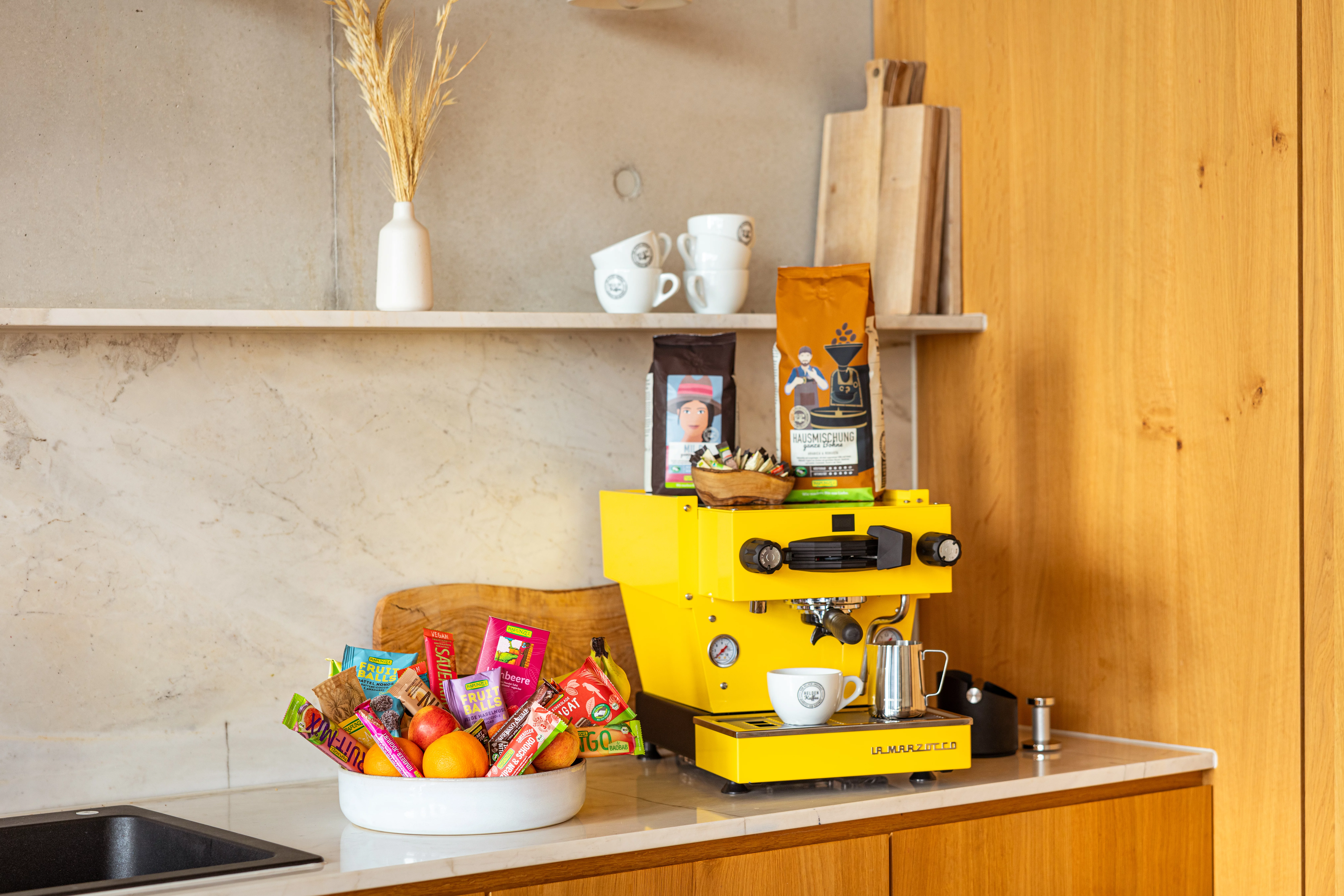Eine Schüssel voller Snacks von Rapunzel. Fruchtschnitten, Fruit Balls & Schokolade. Dahinter steht eine gelbe Kaffeemaschine. 