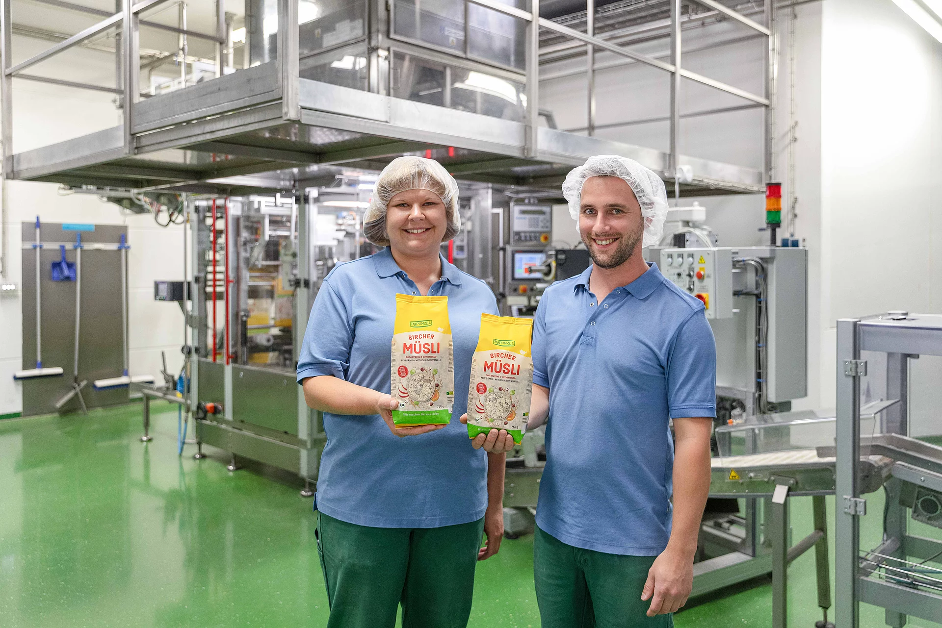 Zwei Mitarbeiter aus der Rapunzel Produktion halten je eine Packung Müsli in die Kamera, im Hintergrund die Müslianlage.