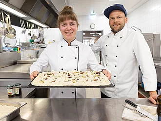 Rapunzel Zutaten und Produkte für Hotels, Gastronomie und Verarbeiter. Rapunzel Köchin präsentiert Focaccia auf Backblech, neben ihr ein Koch, dahinter die Großküche