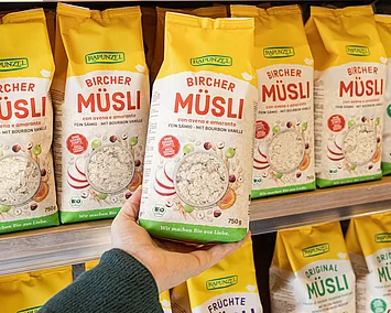 Nahaufnahme von Bircher-Müsli-Packungen im Regal, Hand greift sich eine Packung.