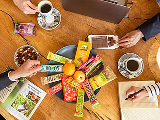 Verschiedene Rapunzel Snacks in einer Schüssel verteilt auf einem Tisch. Mehrere Menschen greifen nach den Snacks. 