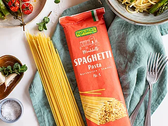 Rapunzel Bio-Spaghetti in Originalverpackung und lose auf Küchentuch, angerichtetes Nudelgericht mit Gemüse drumherum. Rapunzel Bio-Spaghetti werden sowohl in der Originalverpackung als auch lose auf einem Küchentuch präsentiert. Drumherum ist ein angerichtetes Nudelgericht mit Gemüse dekorativ angerichtet.