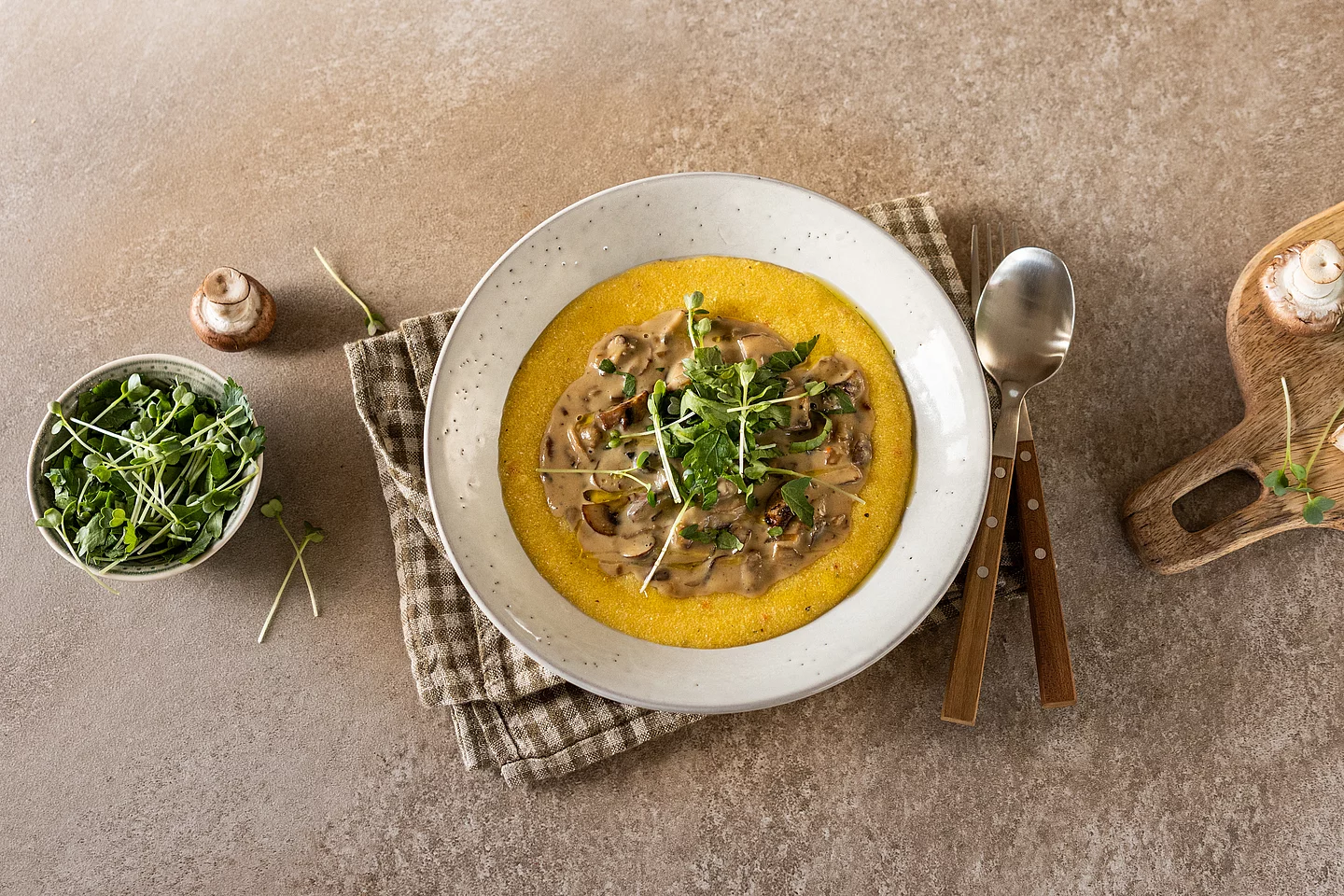 Cremige Polenta mit herzhaftem Pilz-Ragout. Eine Draufsicht auf weißen Teller mit leuchtend gelber, cremiger Polenta, in deren Mitte ein dunkles Pilz-Ragout in einer Sahnesauce angerichtet ist. Garniert mit frischen grünen Sprossen. Der Teller steht auf einer braun-weiß karierten Stoffserviette auf einer rustikalen Steinfläche. Daneben liegen ein Löffel und eine Gabel mit Holzgriffen sowie ein kleines Schälchen mit weiteren Sprossen und frischen Champignons.