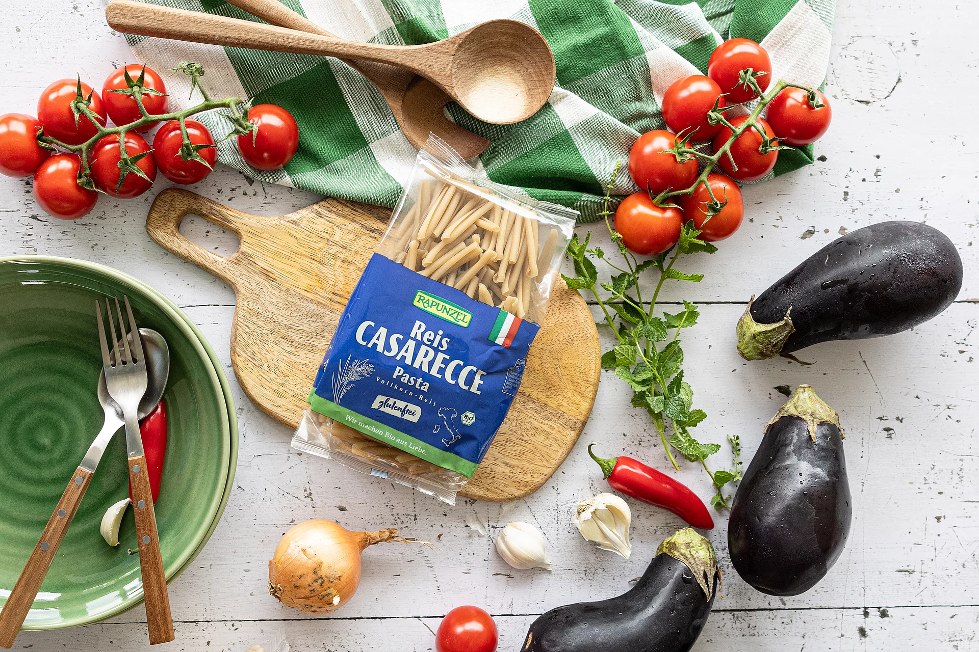 Packung Reis Casarecce Pasta glutenfrei auf einem Schneidebrett, umgeben von Tomaten, Aubergine, Chilischoten, Knoblauch, Minze, Zwiebel, einem grün-weißen Küchentuch, Holzlöffel sowie einer grünen Schüssel mit Gabel und Löffel.