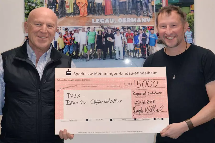 Joseph Wilhelm, Gründer und Gesellschafter von Rapunzel Naturkost (links) übergibt den Scheck in Höhe von 5.000 Euro an Heinz Ratz, Musiker und Initiator von BOK – Büro für Offensivkultur.